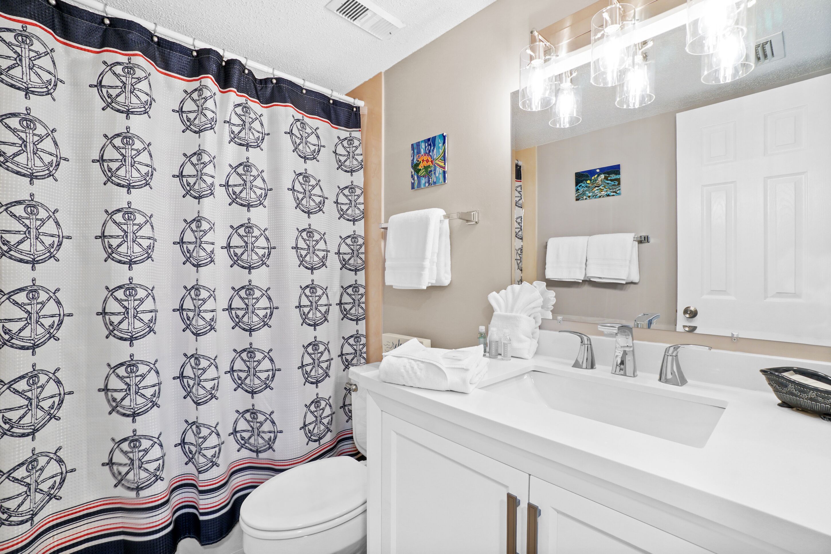 Un baño con cortina de ducha de temática náutica, un lavamanos blanco con espejo y un inodoro blanco.