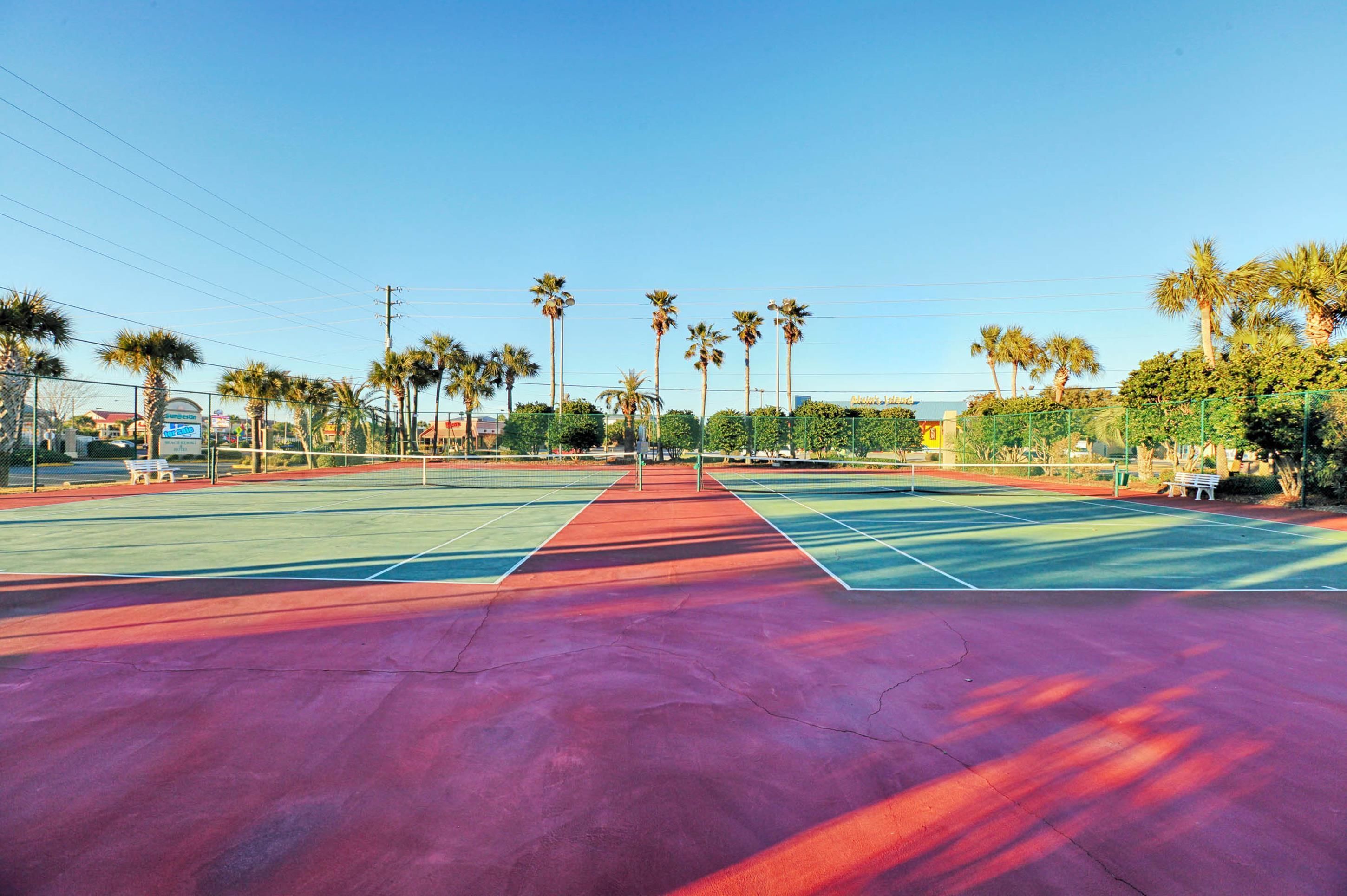 A tennis court with multiple courts, surrounded by palm trees and benches.