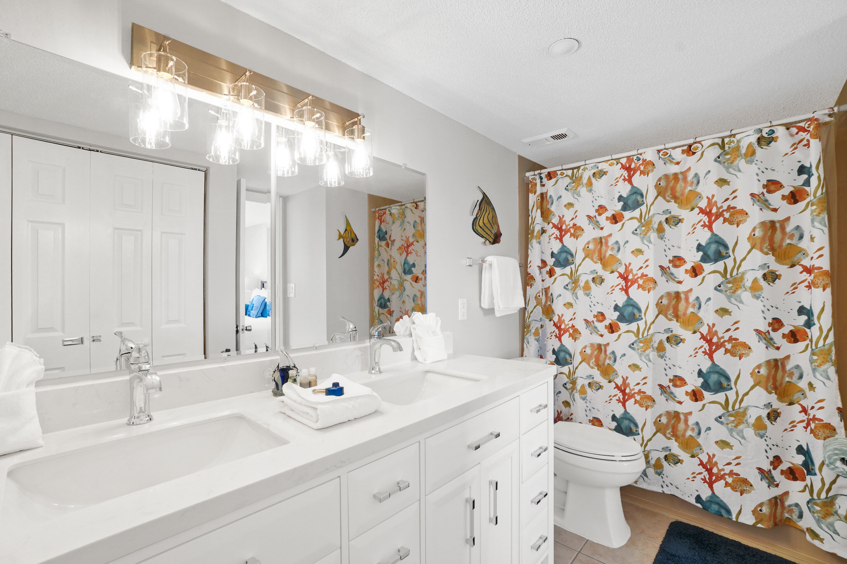 A bathroom with a white vanity, a large mirror, a sink, a toilet, and a shower curtain with a fish pattern.