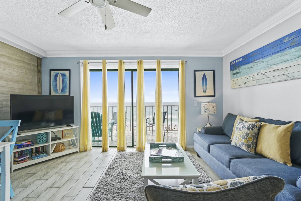 A living room with a TV, sofa, and a view of the beach through large windows.