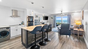 A modern kitchen with a central island, bar stools, a dining area with a view, and a living space with a sofa and lamp. - Summit Beach Resort by Panhandle Getaways (Panama City Beach)