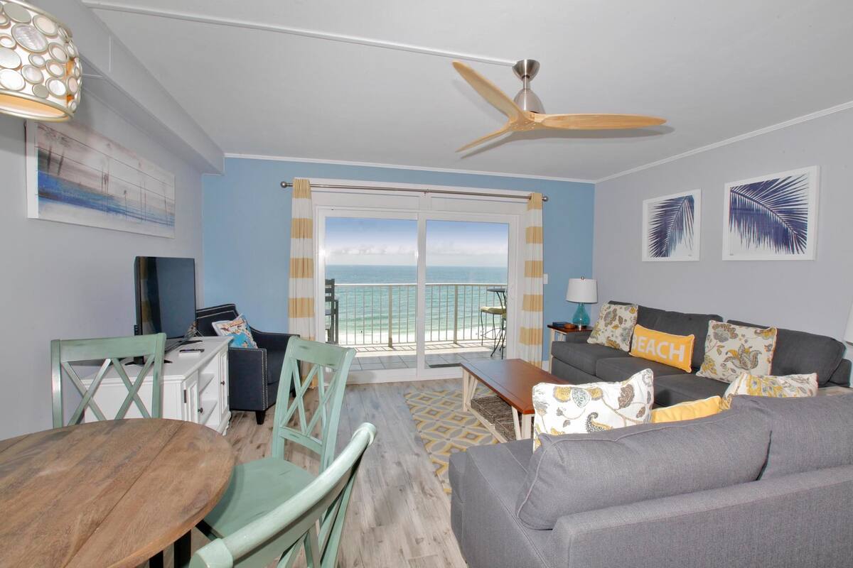 A living room with a view of the ocean, a ceiling fan, and a dining area with a table and chairs.