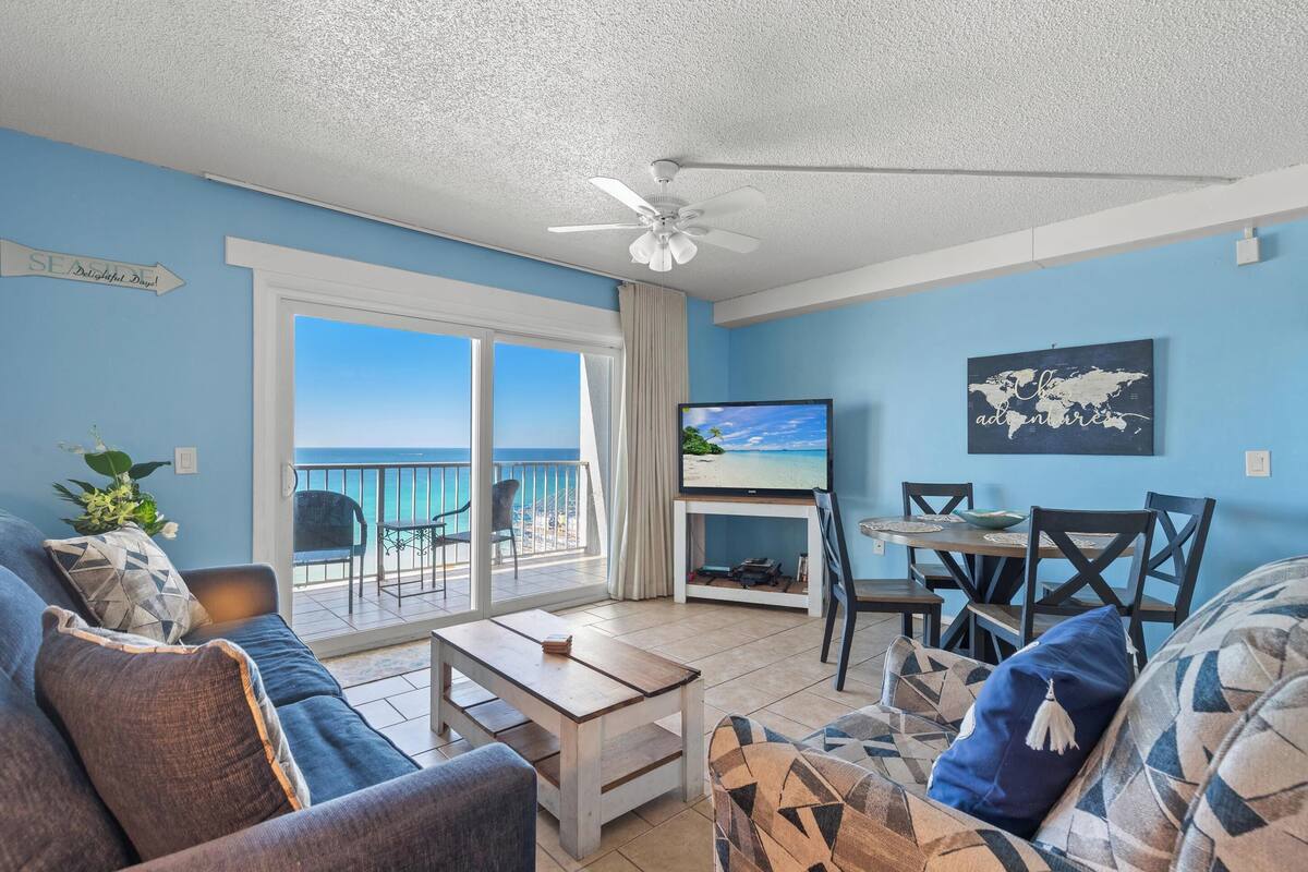 A living room with a sofa, coffee table, TV, and a dining area with chairs and a table. There is a balcony with a view of the ocean.