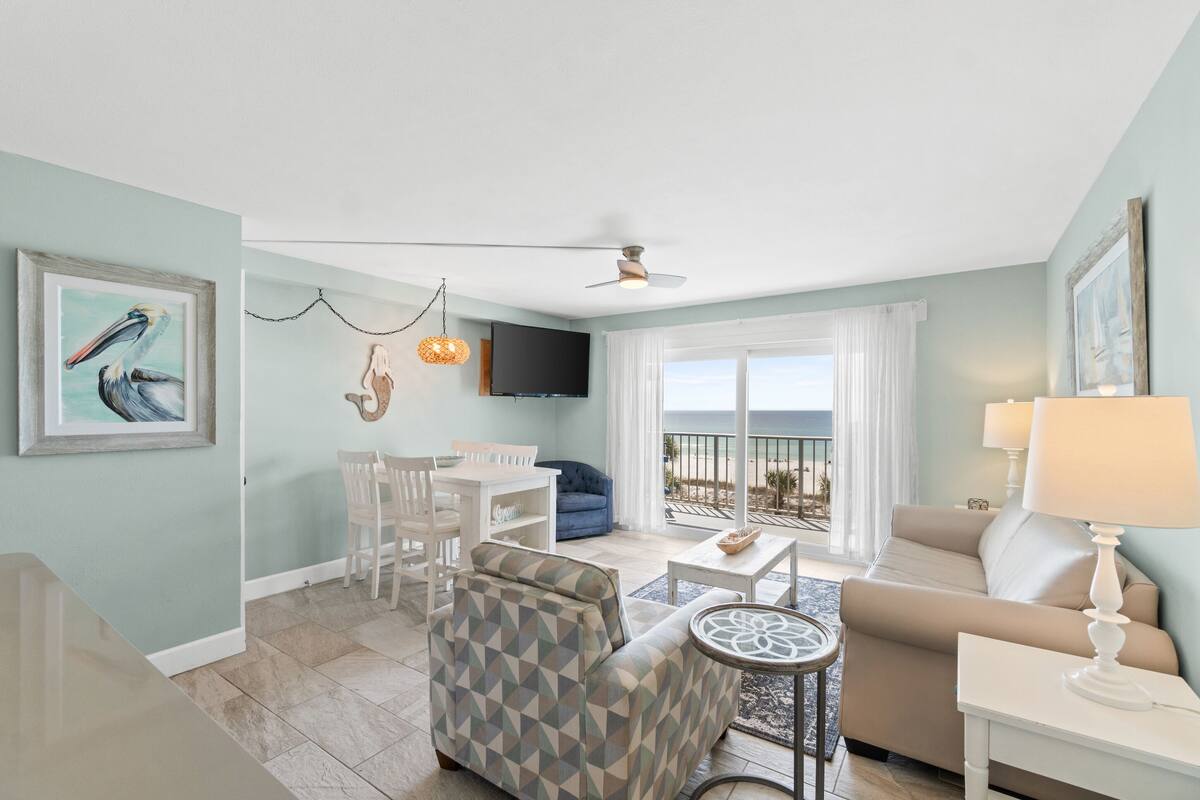 A living room with a sofa, armchair, dining table, and chairs. There is a TV mounted on the wall and a ceiling fan.