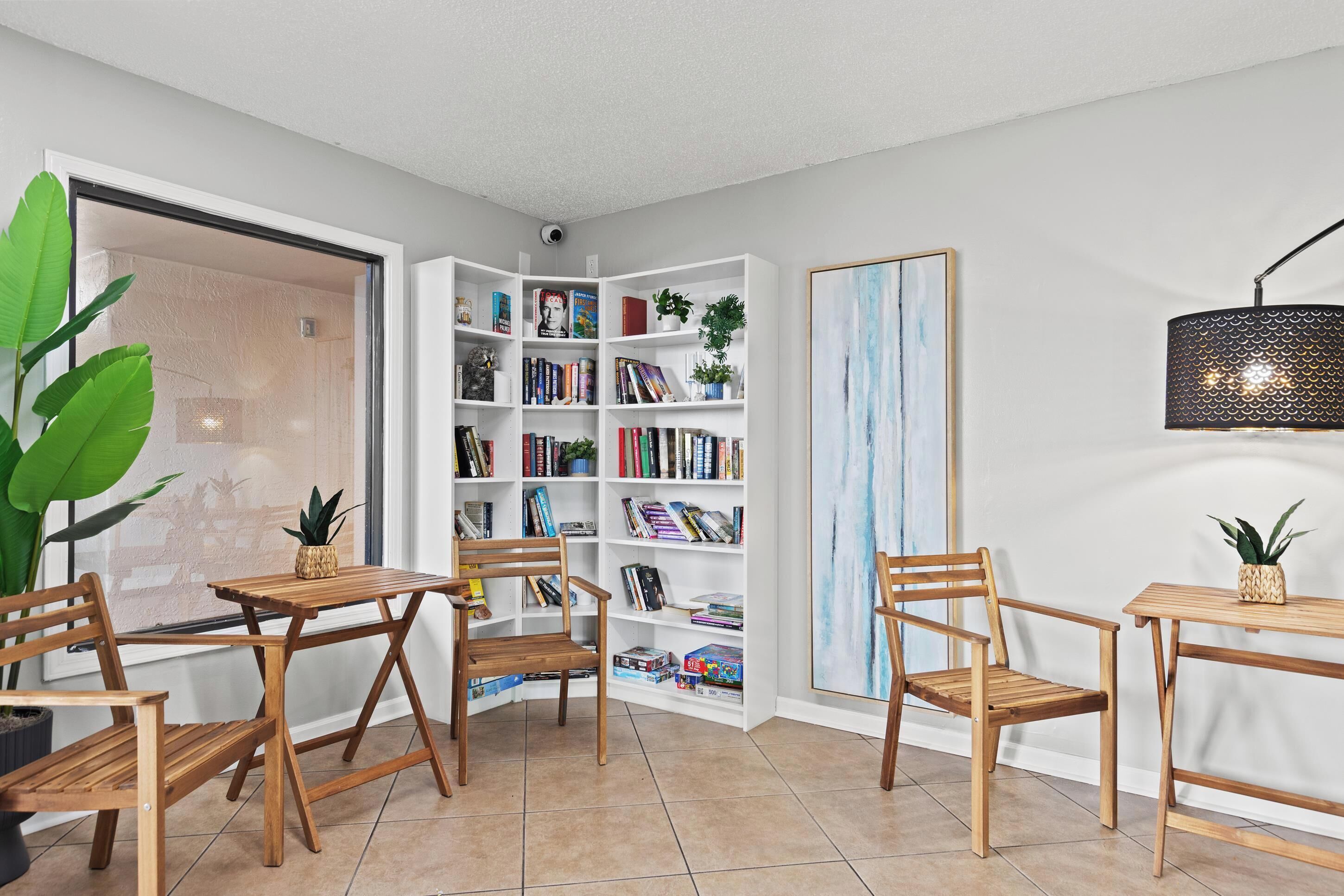 A modern living room with a wooden bookshelf, a dining table, chairs, and a large window.