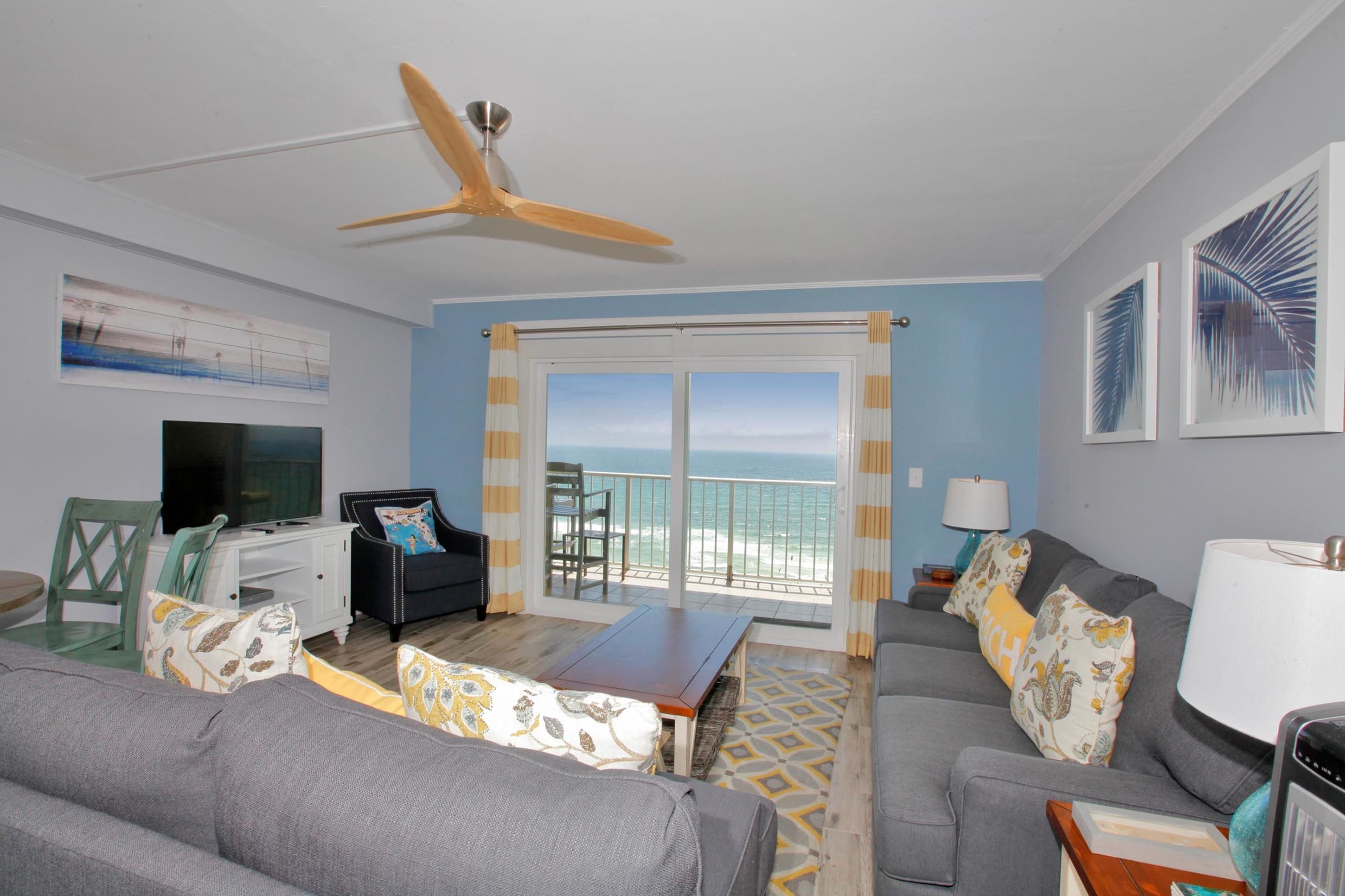 A living room with a ceiling fan, a television, a sofa, a coffee table, and a view of the ocean through a sliding glass door.