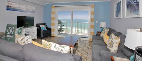 A living room with a ceiling fan, a television, a sofa, a coffee table, and a view of the ocean through a sliding glass door.
