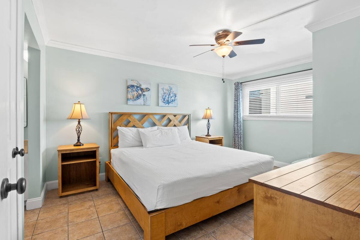 A bedroom with a wooden bed, two bedside tables with lamps, a ceiling fan, and artwork on the wall.