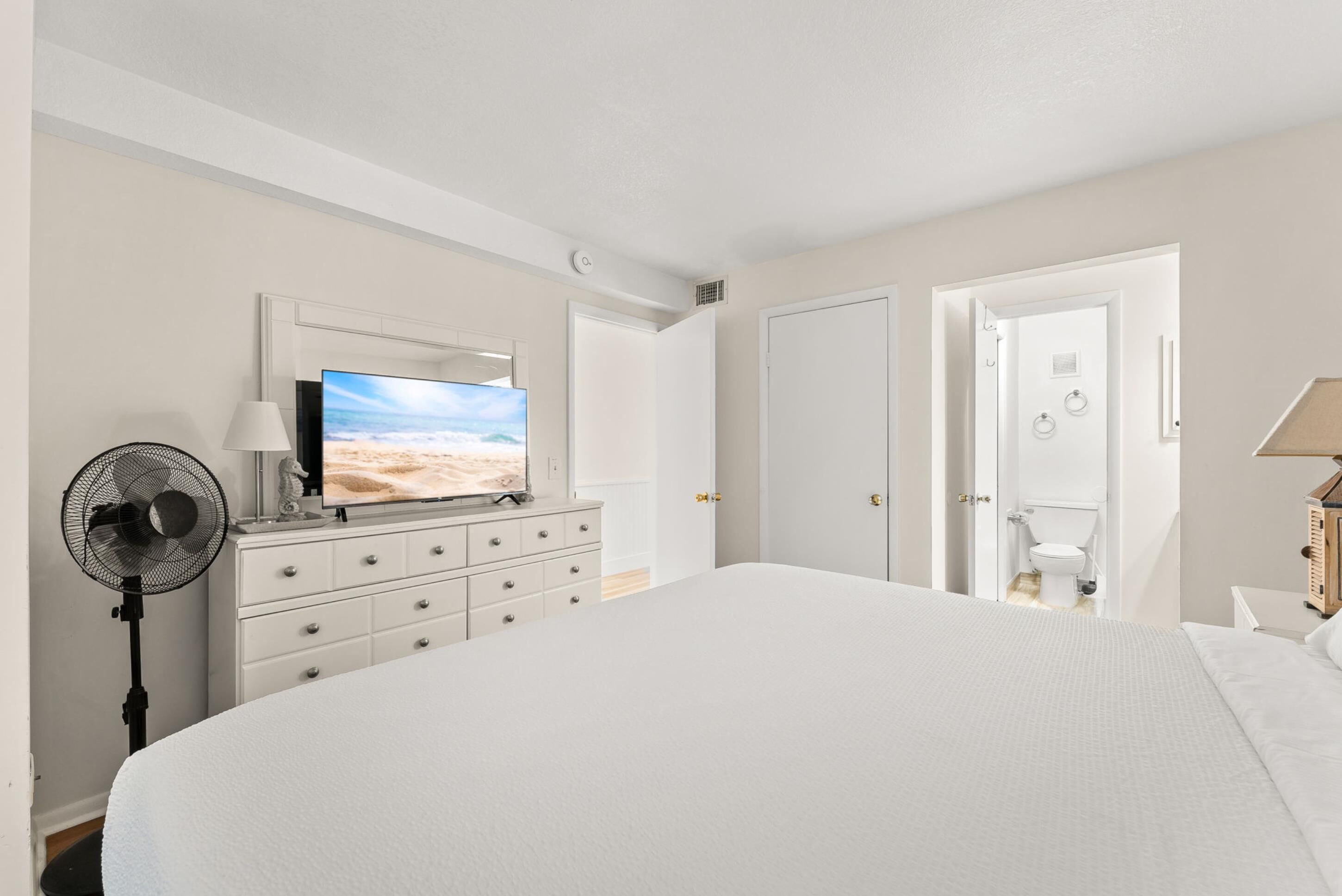 Een slaapkamer met een groot bed, een televisie op een dressoir, een ventilator en een deur naar een badkamer.