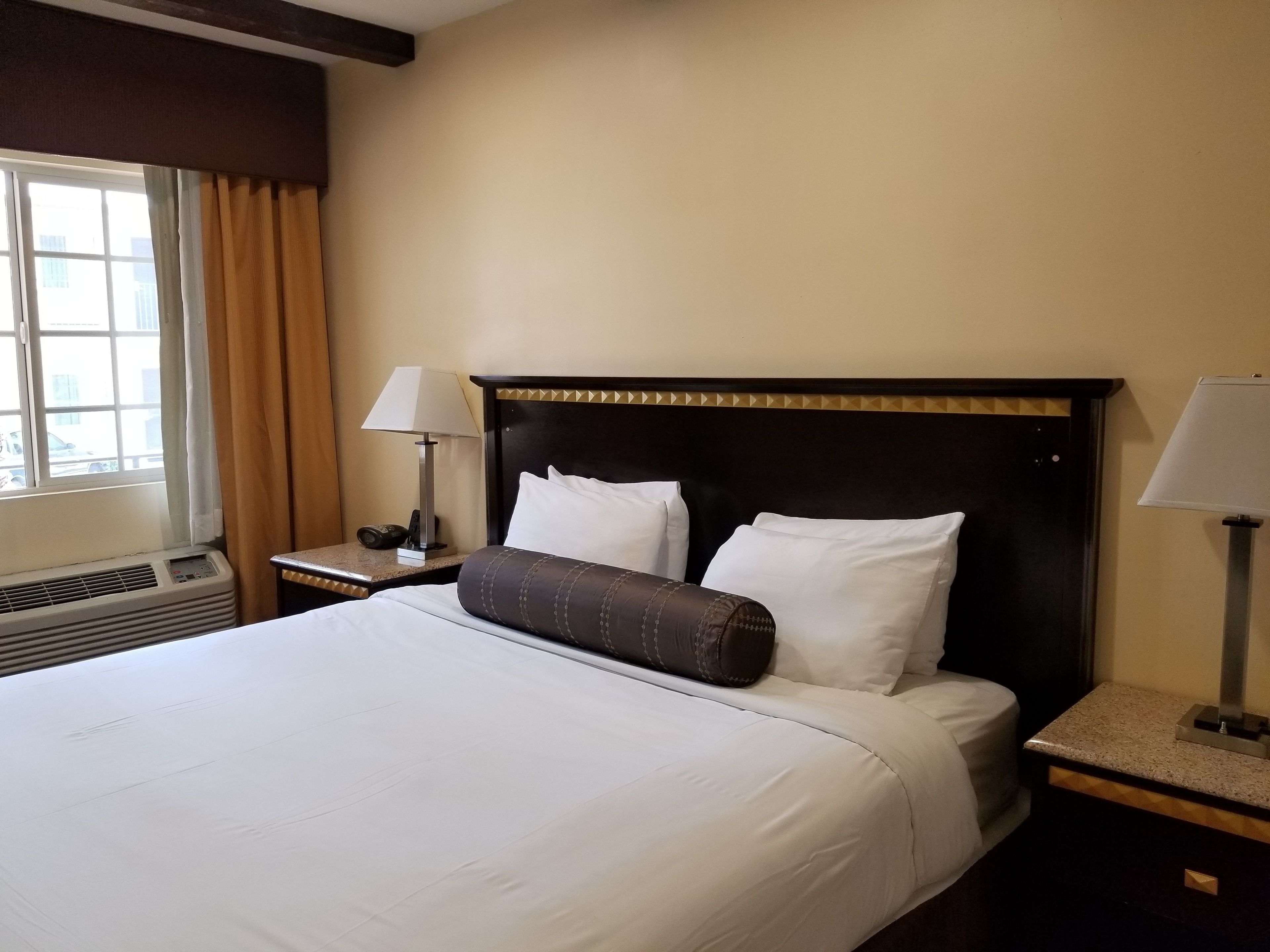 A hotel room with a large bed, two bedside tables with lamps, a window with curtains, and an air conditioning unit.