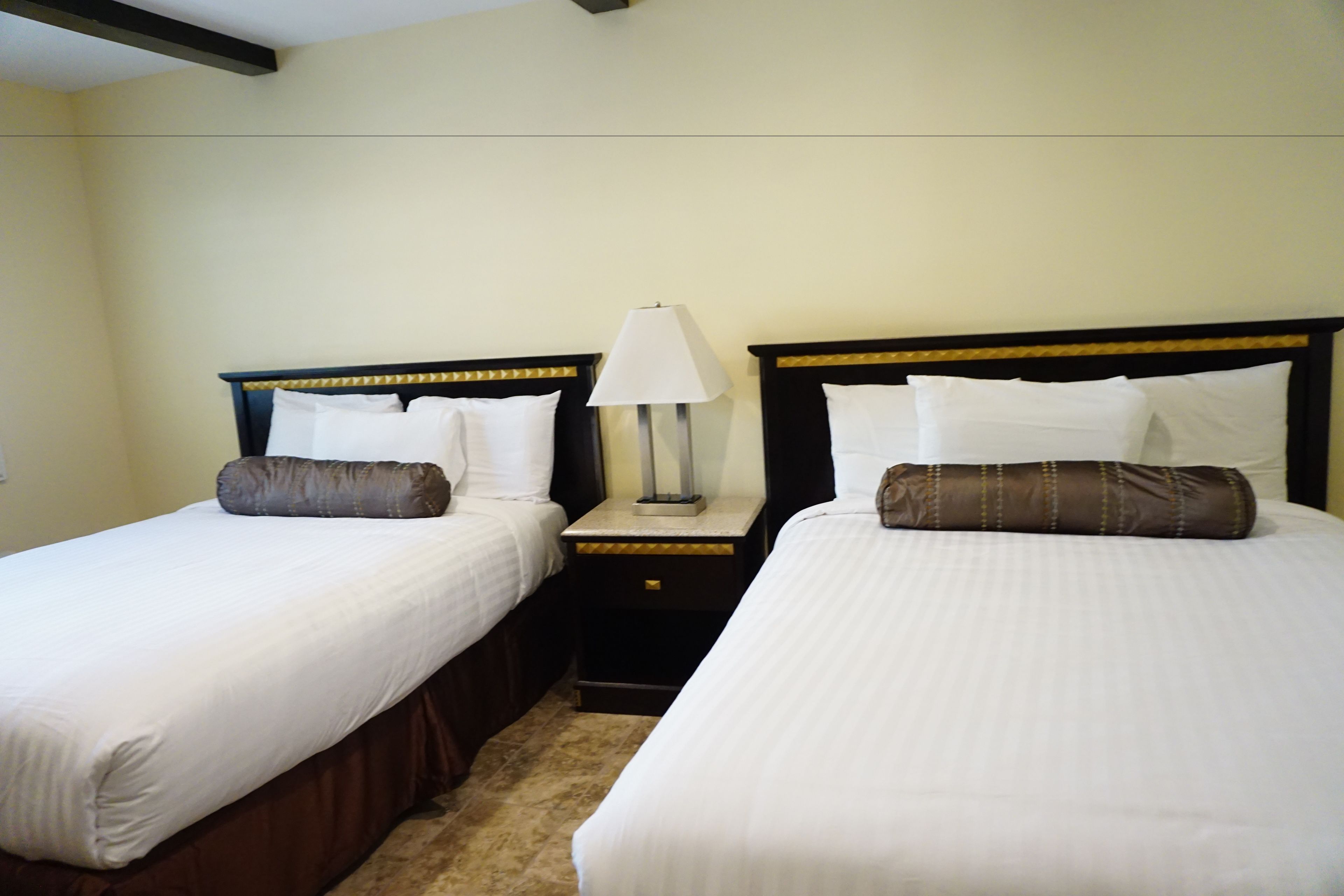 A hotel room with two single beds, each with a pillow and a bedside table with a lamp.