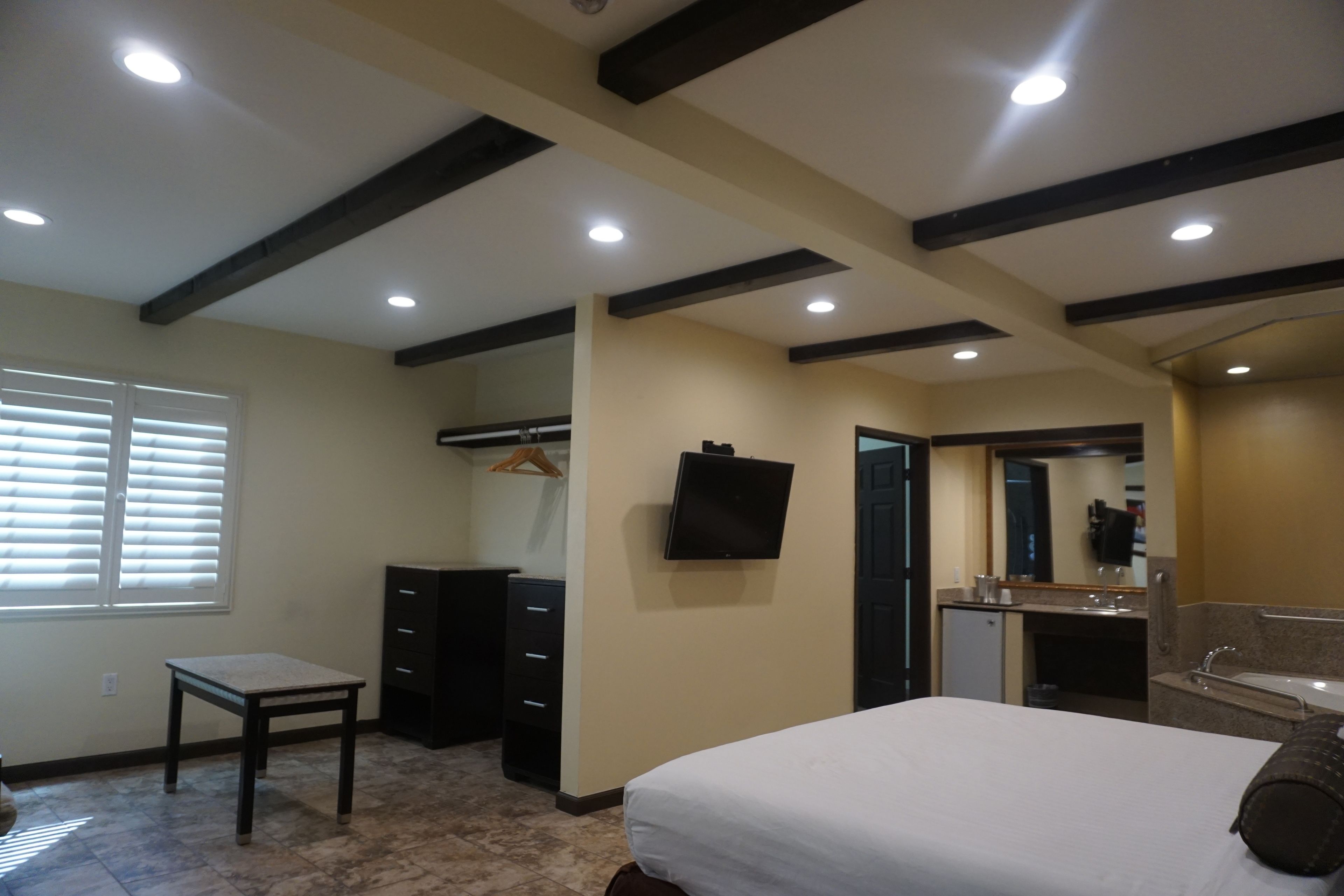 A hotel room with a bed, a desk, a dresser, a TV, a bathroom with a sink and mirror, and a window with blinds.