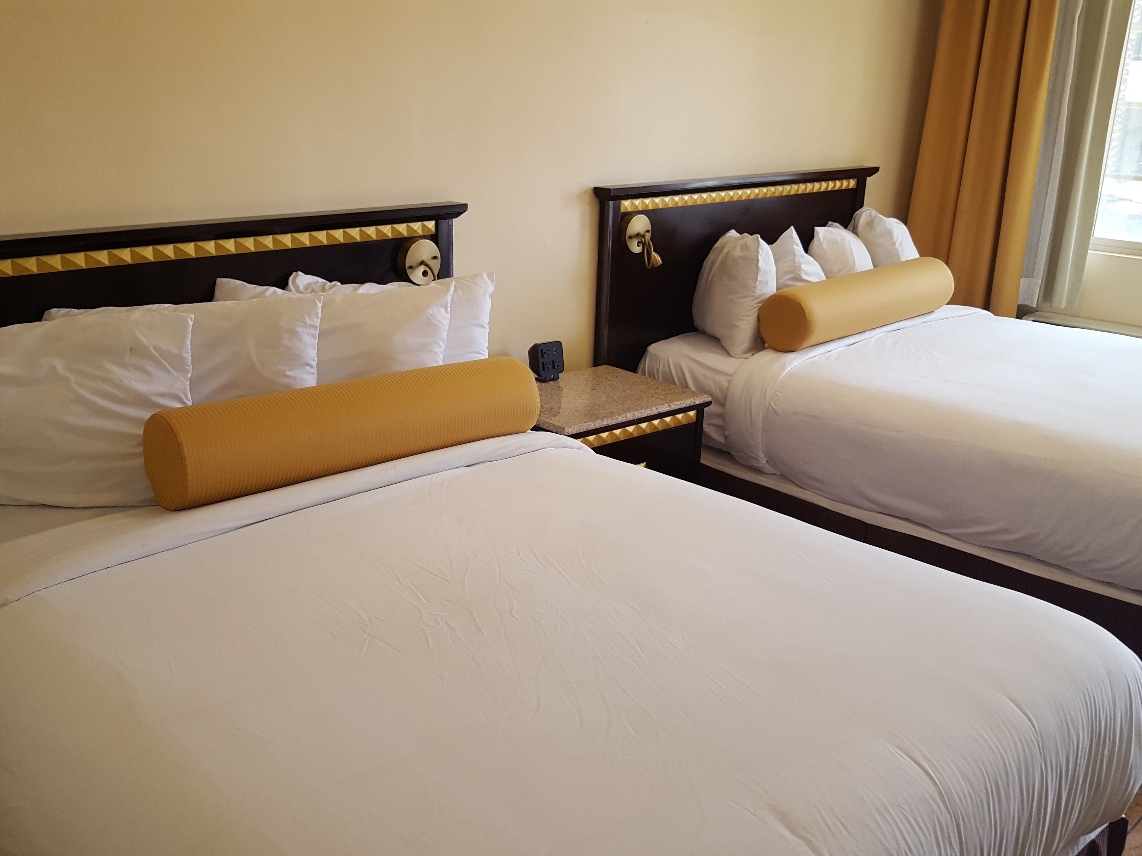 Ein Hotelzimmer mit zwei Betten, jeweils mit weißer Bettwäsche und einem gelben Akzentkissen. Zwischen den Betten befindet sich ein Nachttisch mit einer Lampe und einer Steckdose.