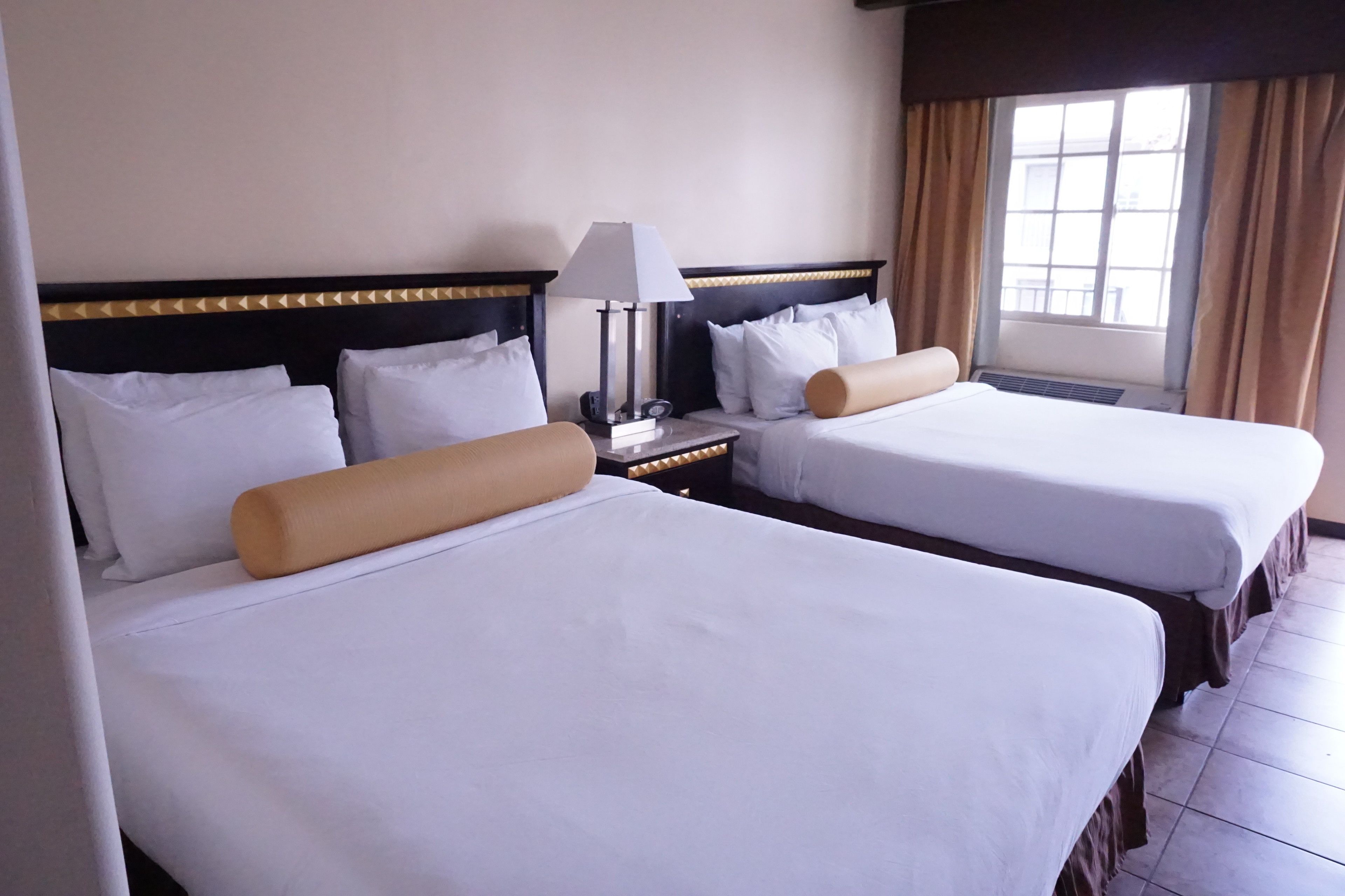 A hotel room with two beds, a nightstand with a lamp, and a window with curtains.