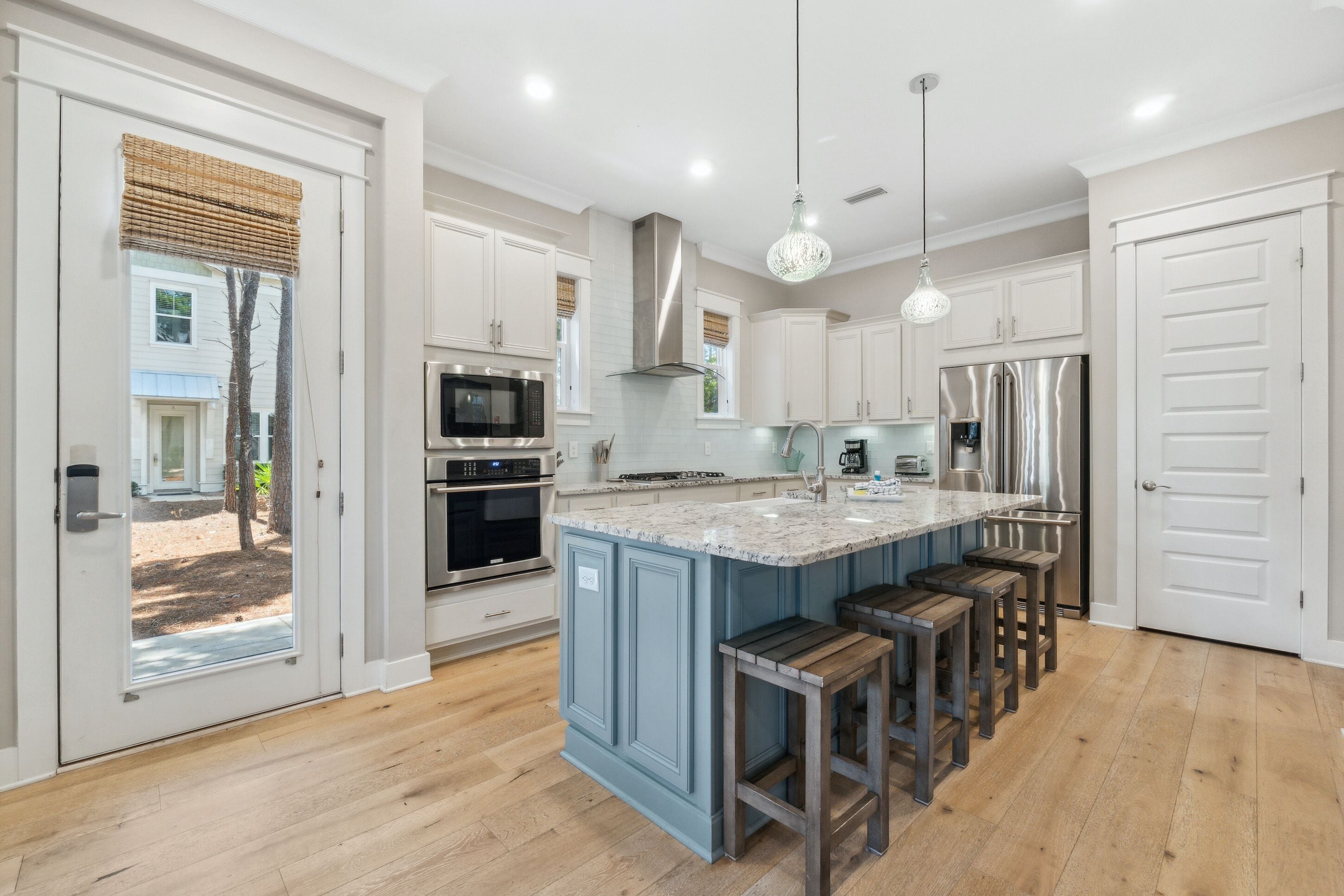 A modern kitchen with white cabinets, a large island with bar stools, stainless steel appliances, and a view through the glass door.