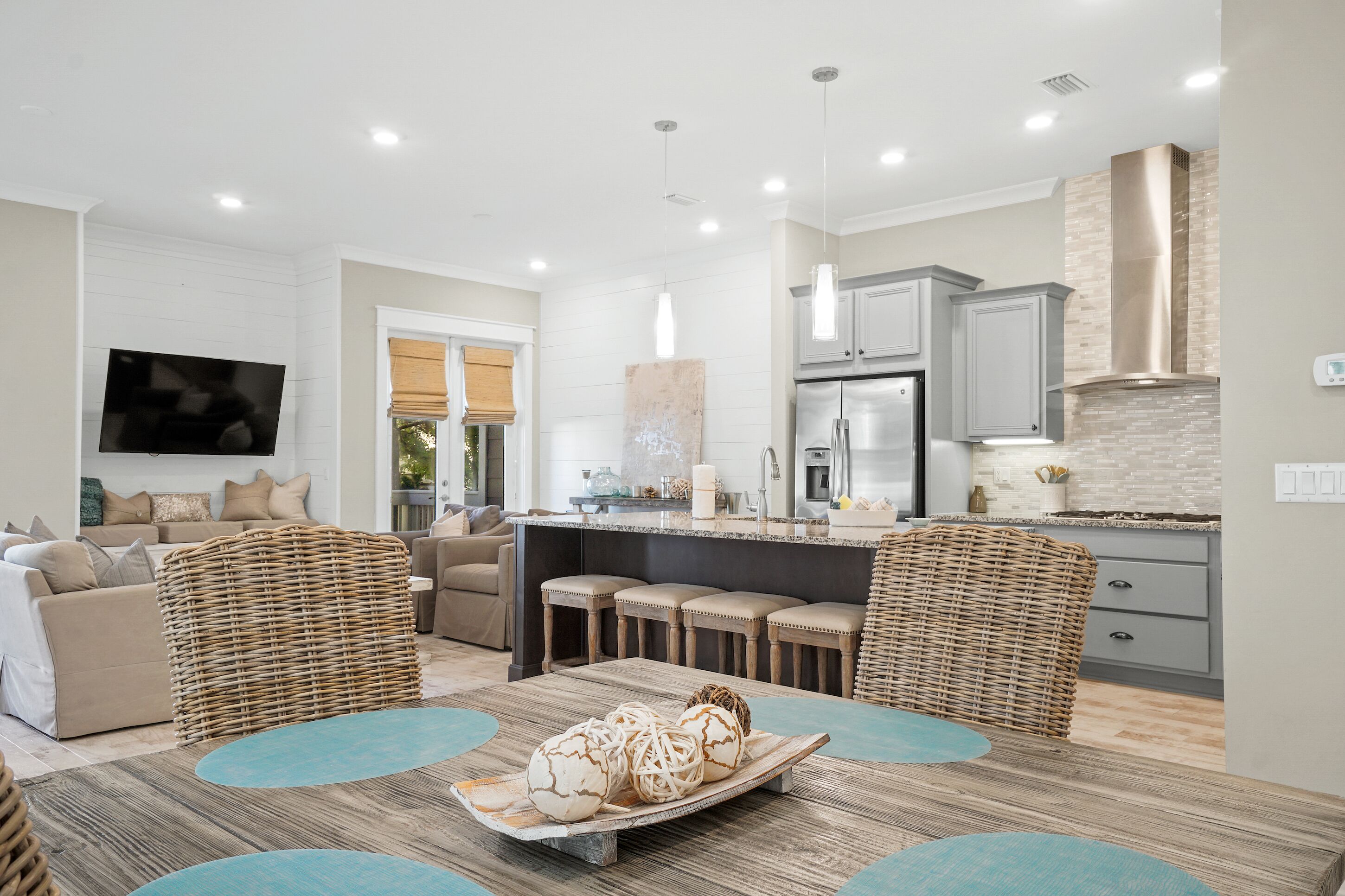 A modern living room with a large TV, comfortable seating, a kitchen with grey cabinets, and a central island.