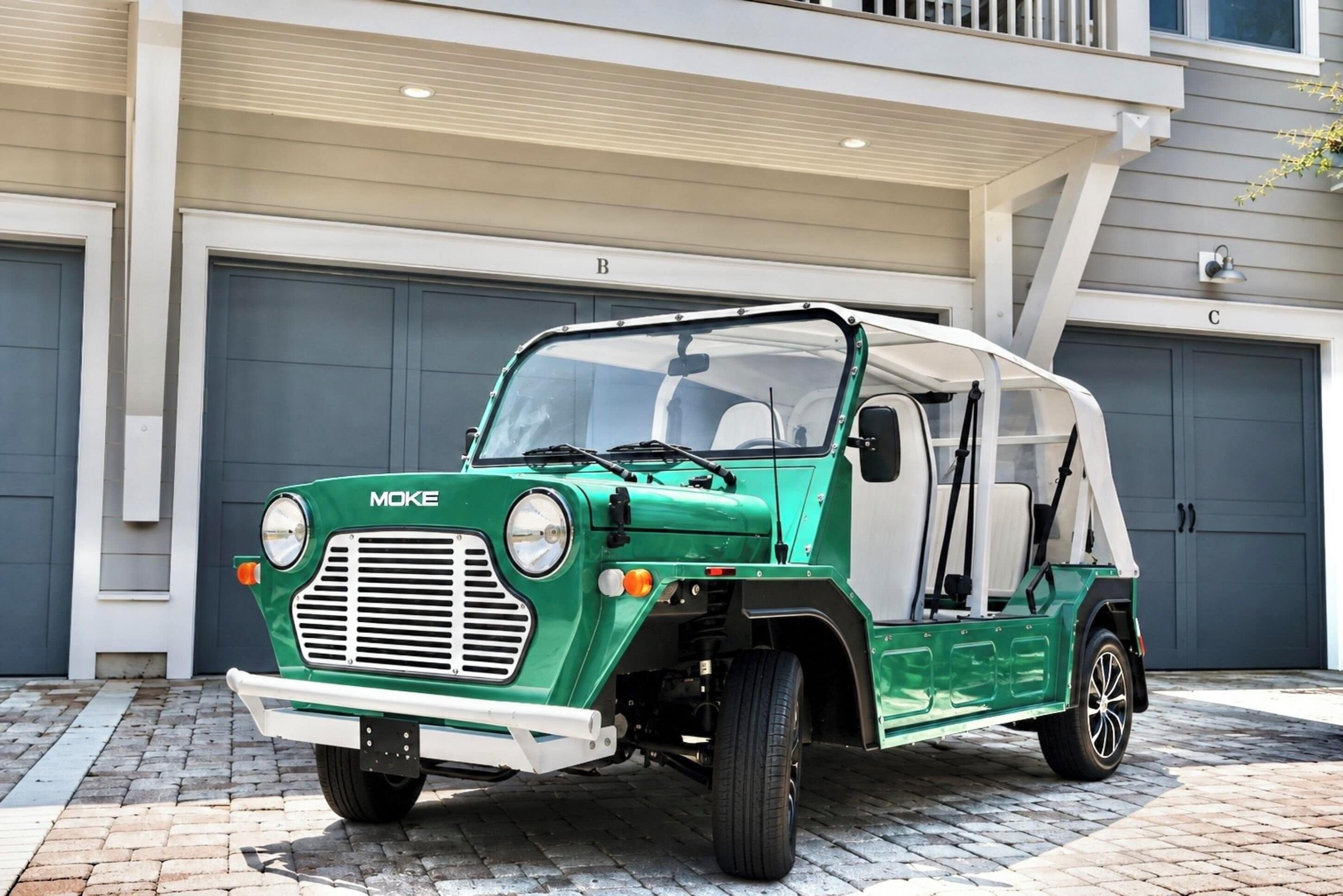 Um veículo MOKE verde estacionado em frente a um edifício com portas de garagem identificadas com as letras B e C.