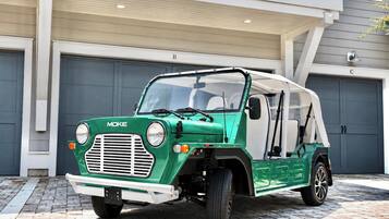 Um veículo MOKE verde estacionado em frente a um edifício com portas de garagem identificadas com as letras B e C.