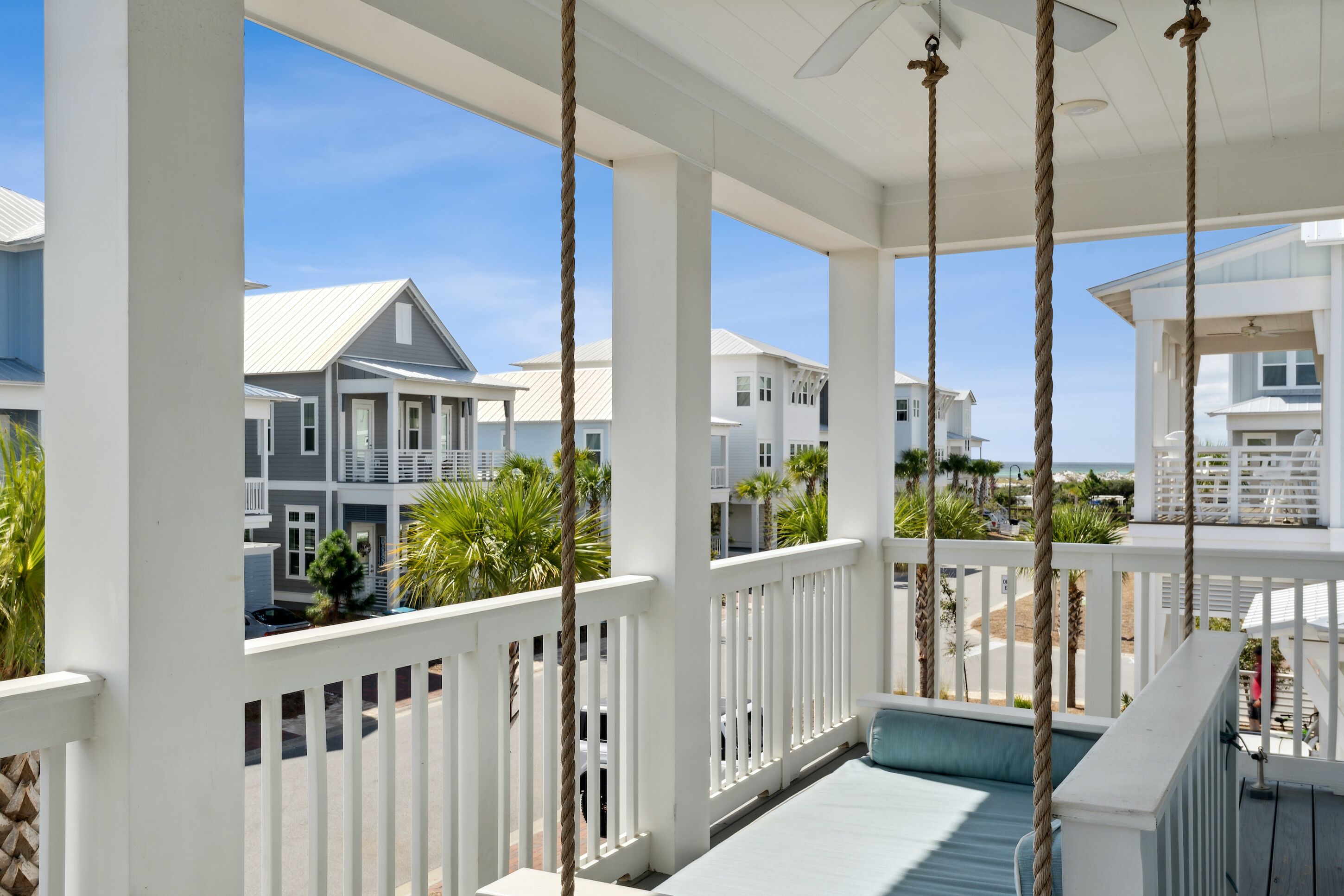 A covered porch with white railings, a ceiling fan, and a view of neighboring houses and palm trees.