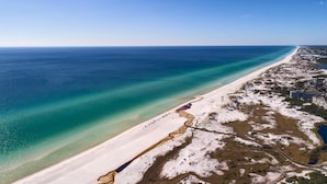 Vista aérea de una zona costera con una extensa playa de arena, un océano azul cristalino y algunos edificios a lo largo de la costa.