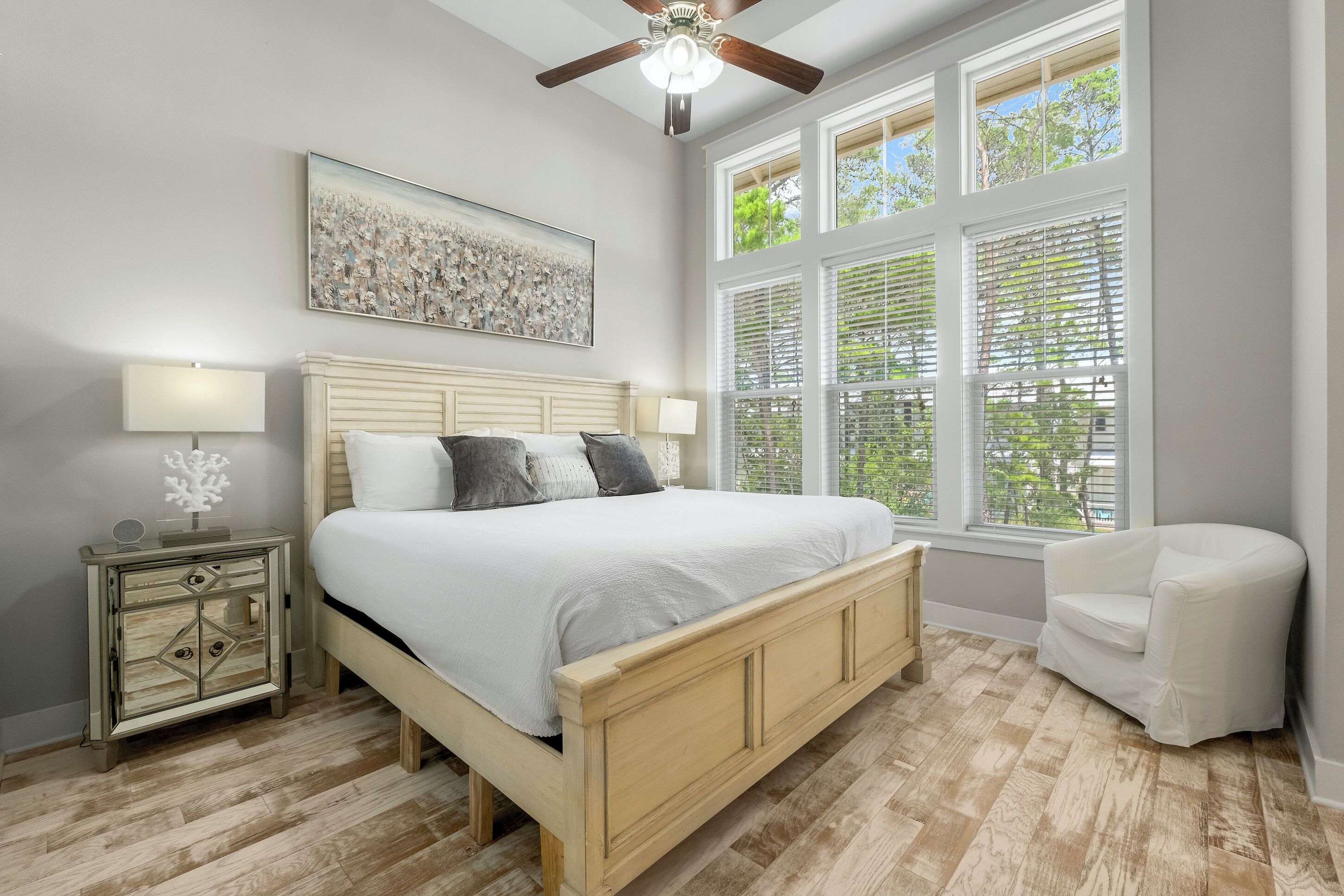 A bedroom with a wooden bed, a nightstand with a lamp, a chair, and a large window with blinds.