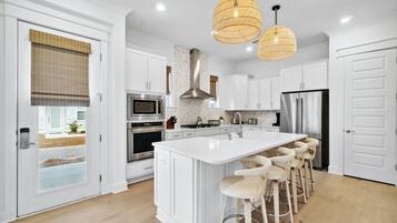 Una cocina moderna con armarios blancos, una isla grande con taburetes de bar, electrodomésticos de acero inoxidable y dos lámparas colgantes.