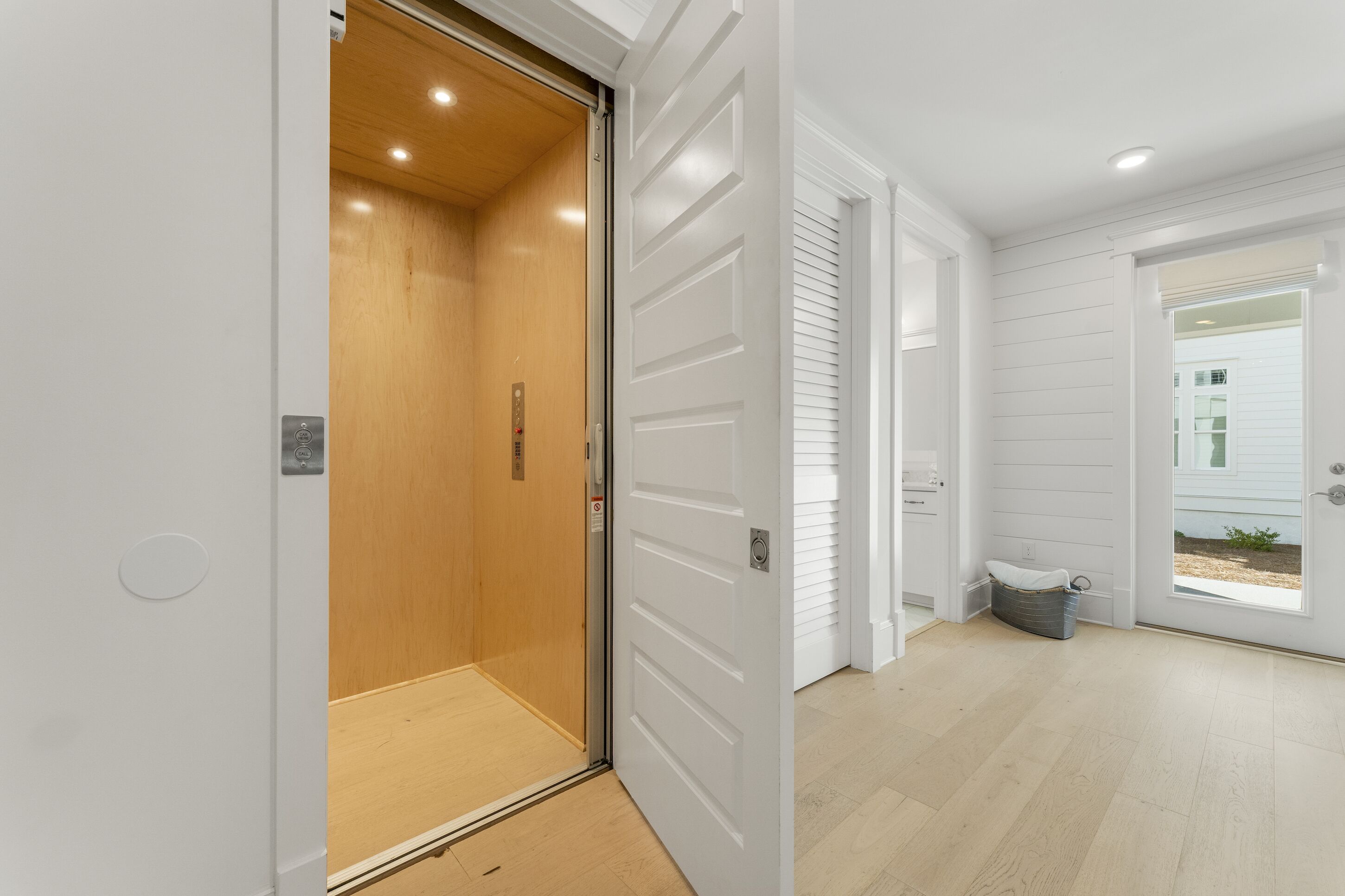 A modern interior with a wooden elevator, white doors, and a view of a house through a glass door.