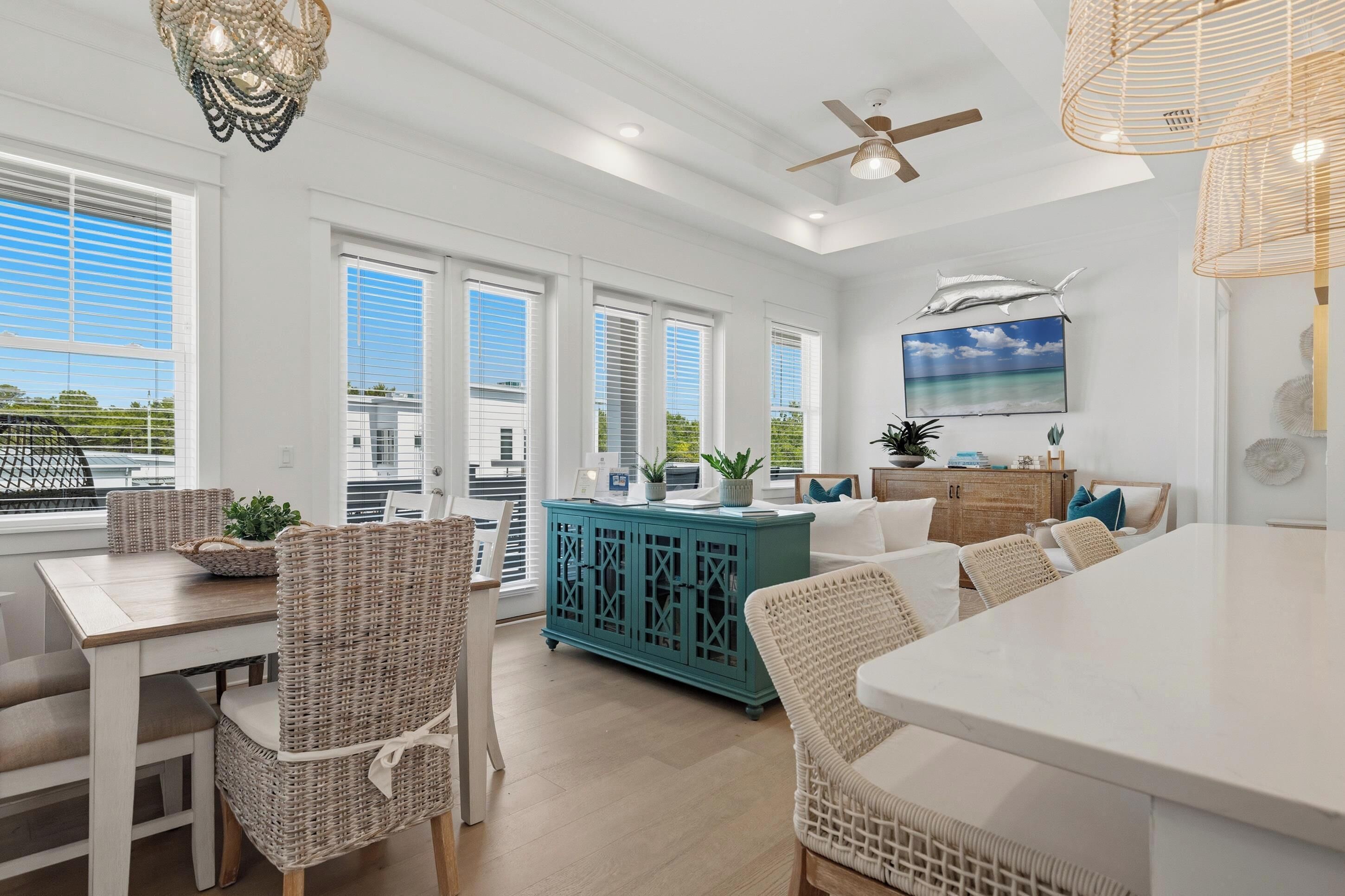 A bright, airy living room with a large dining table, wicker furniture, a teal cabinet, and a ceiling fan.