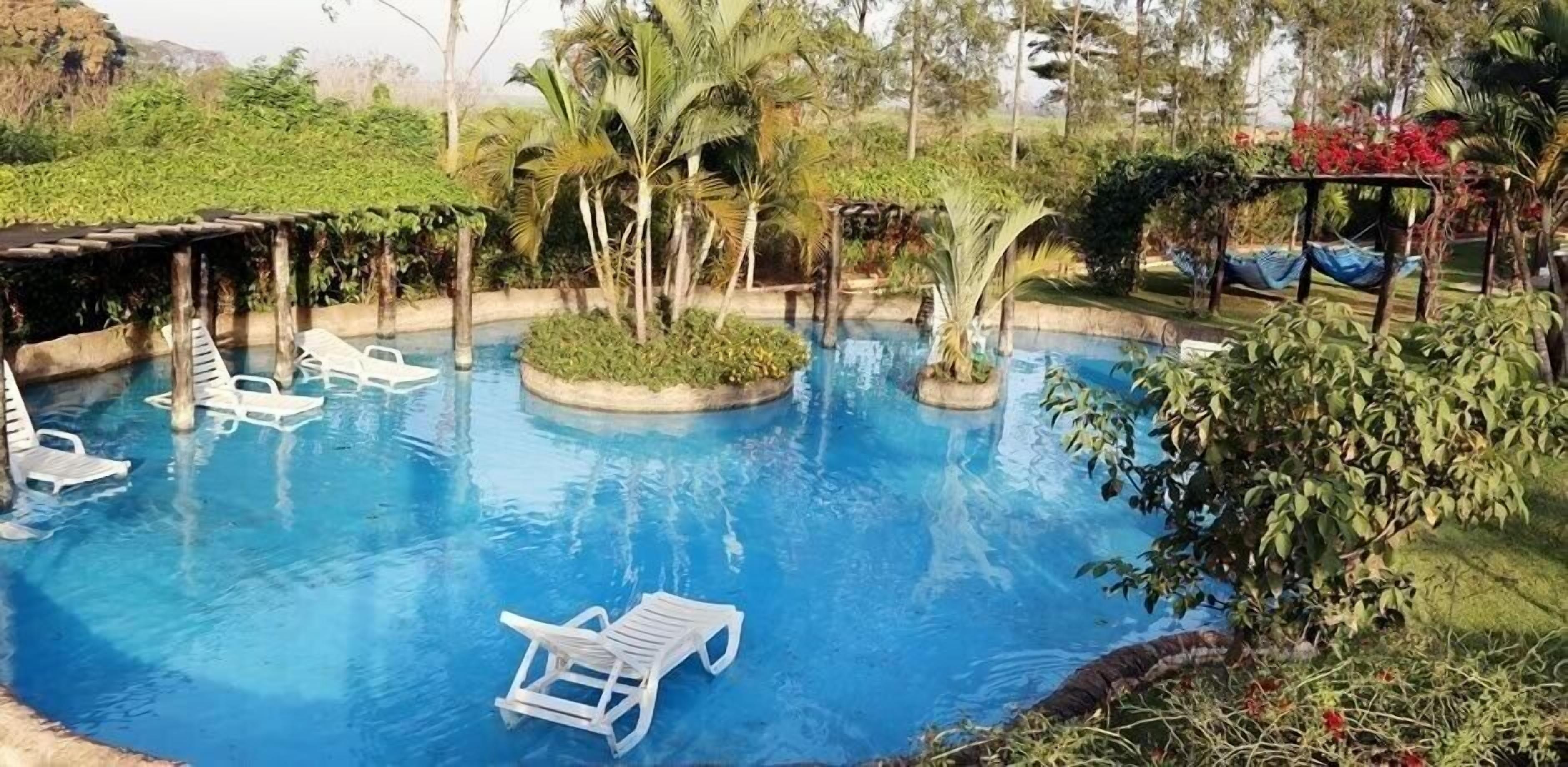 Piscina com cadeiras de descanso brancas, rodeada por vegetação e um pergolado.
