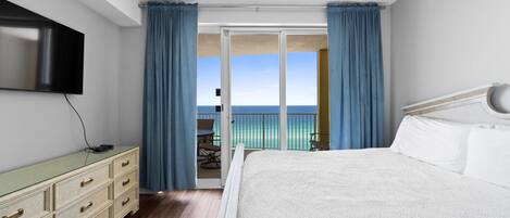 Un dormitorio con una cama grande, un televisor de pantalla plana en la pared, un aparador y un balcón con vista al océano.