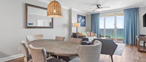Un comedor con una mesa redonda de madera y sillas tapizadas, un ventilador de techo y un ventanal con cortinas.