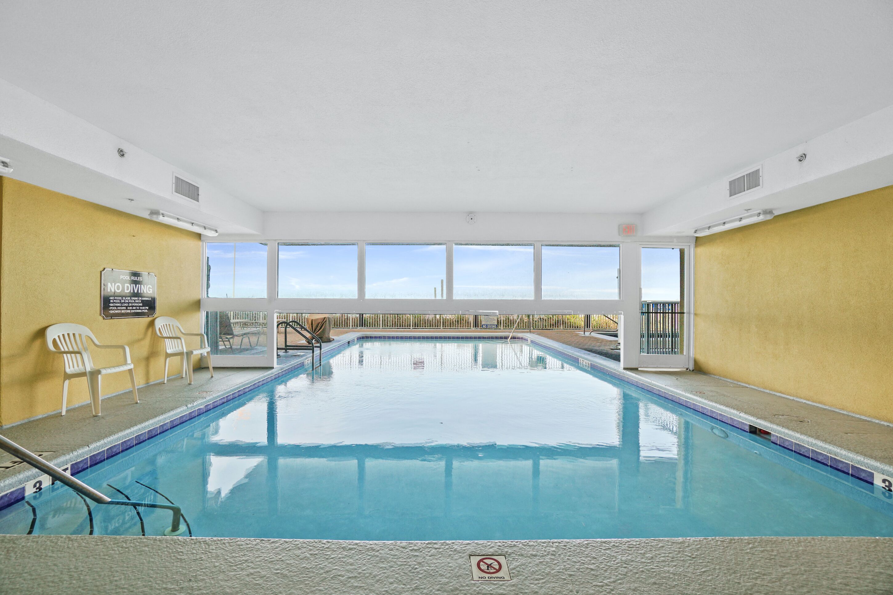 Une piscine intérieure à l’eau d’un bleu limpide, agrémentée d’un panneau indiquant « Interdiction de plonger », et offrant une vue sur le ciel grâce à de grandes fenêtres.