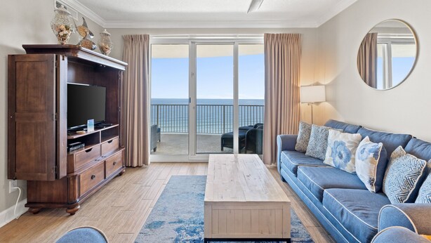 A living room with a large TV, a wooden cabinet, a blue sofa with patterned cushions, a coffee table, and a view of the ocean through a sliding glass door. - Emerald Isle Beach Resort by Panhandle Getaways (Panama City Beach)