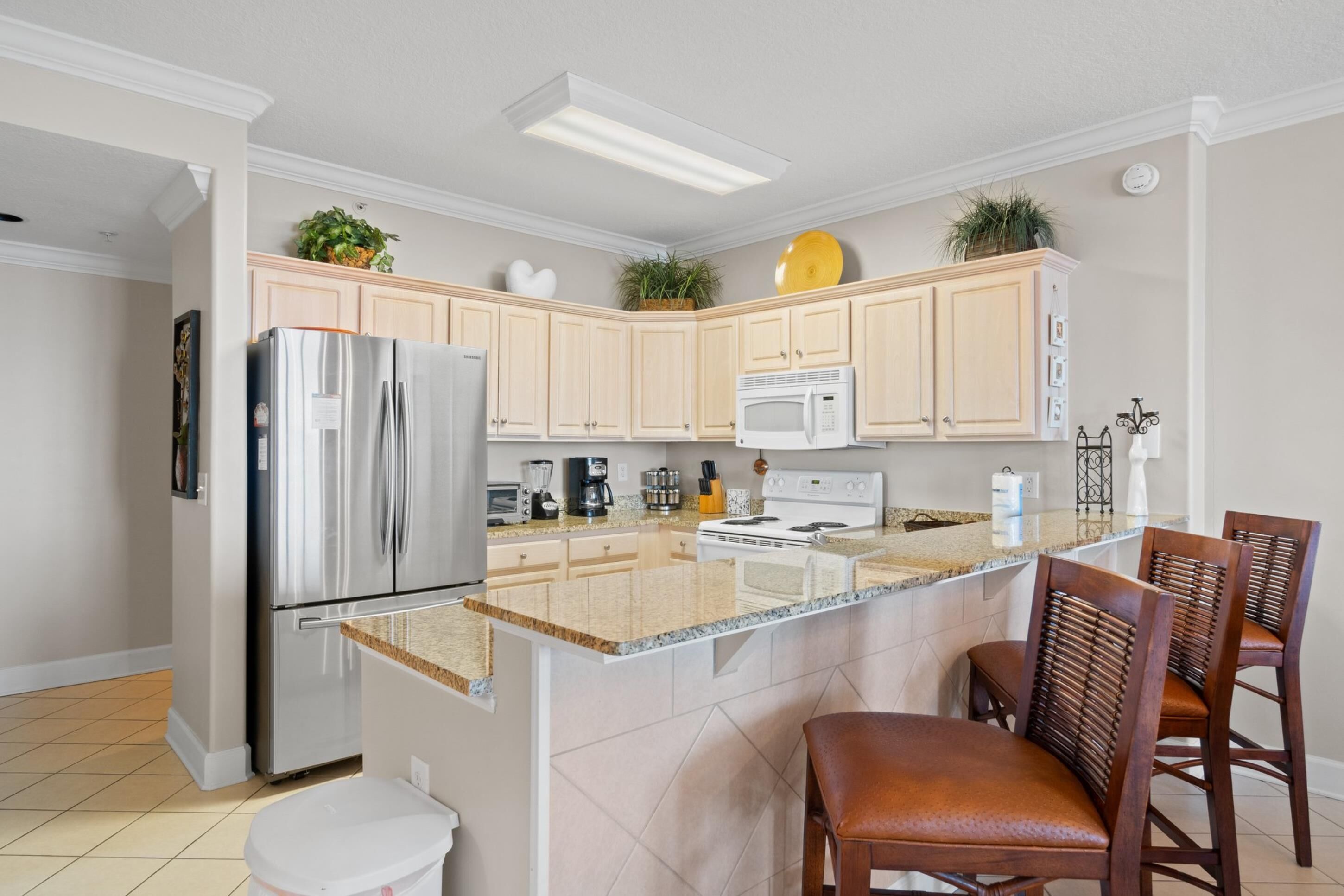 A modern kitchen with a stainless steel refrigerator, a microwave, a stove, and a granite countertop. There are wooden cabinets and a small island with two wooden chairs.