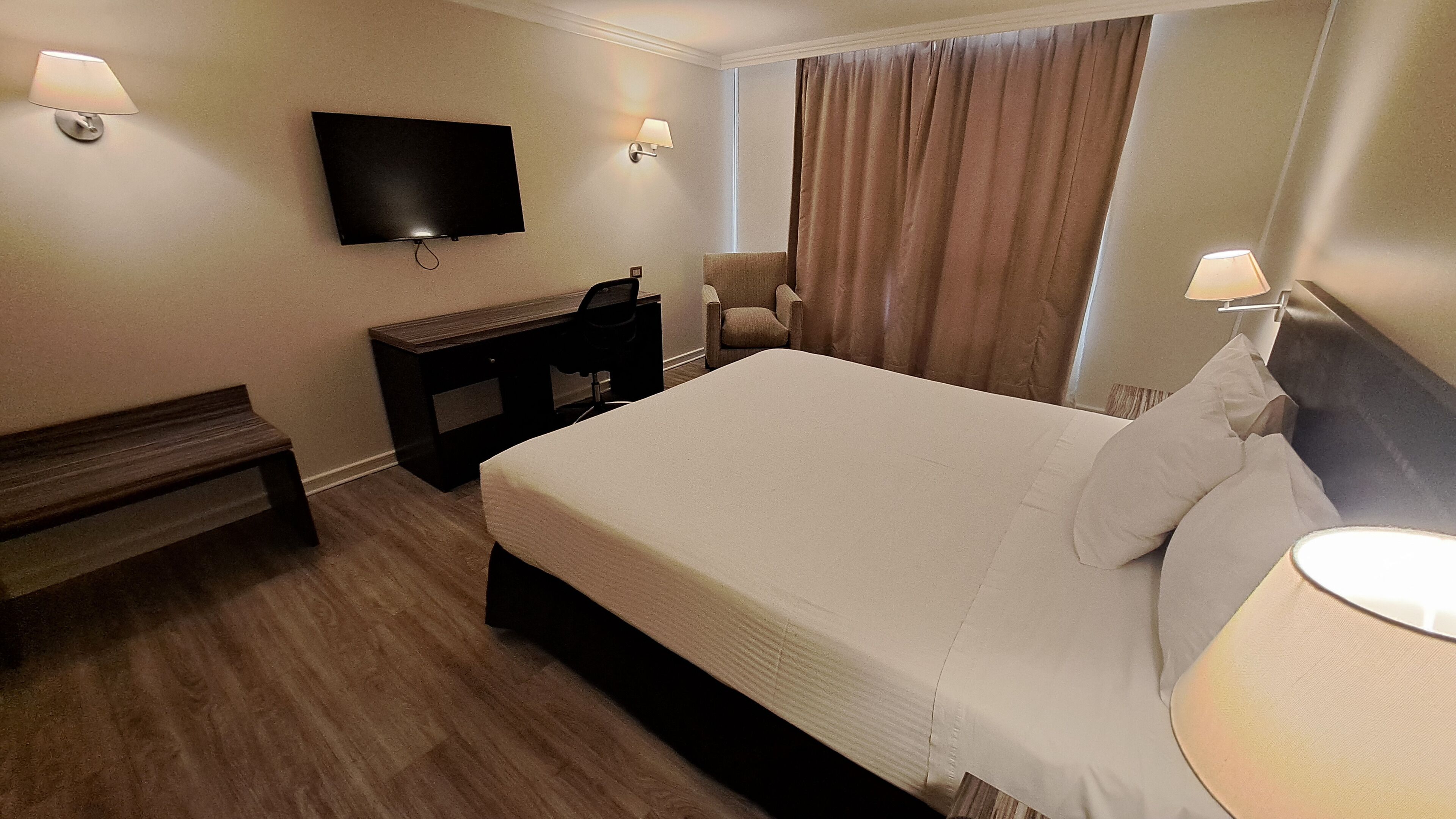 Una habitación de hotel con una cama grande, un escritorio, una silla, un televisor montado en la pared y una ventana con cortinas.
