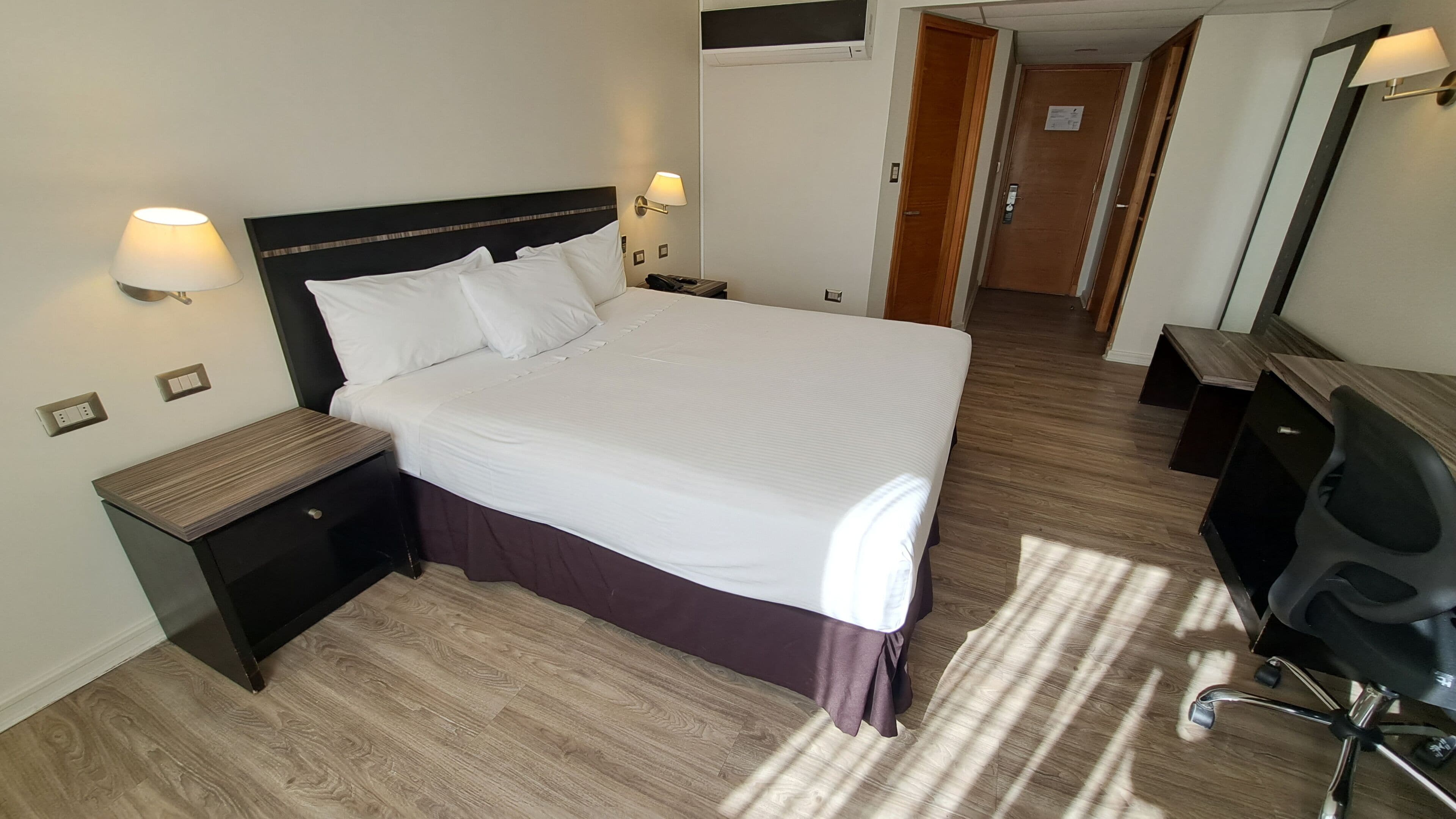 Una camera d'albergo con un letto grande, comodini, una scrivania con una sedia, uno specchio e una porta che dà su un'altra stanza.