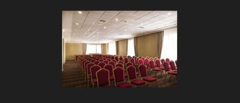 A conference room with rows of red chairs, a carpeted floor, and a high ceiling with recessed lighting.