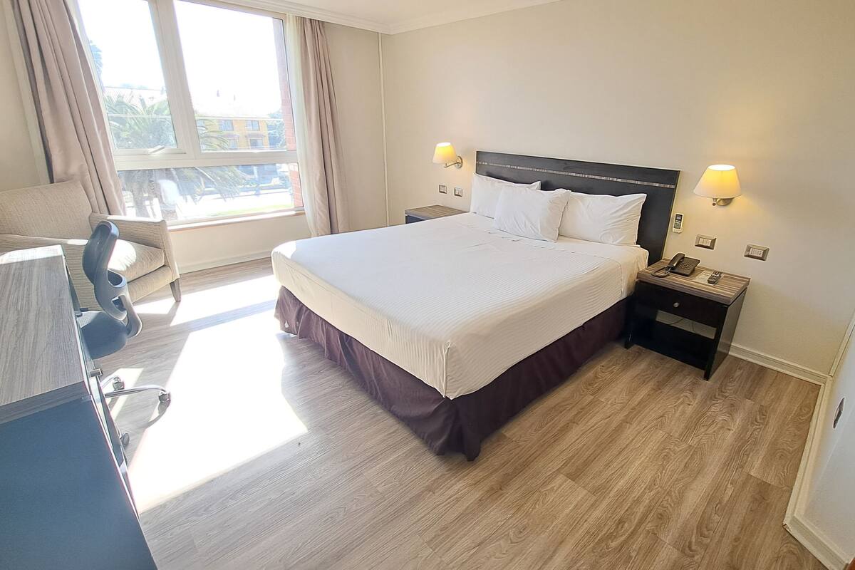 A hotel room with a large bed, two bedside tables with lamps, a desk with a chair, and a window with curtains.