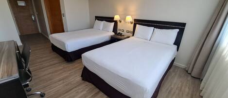 Quarto de hotel com duas camas de solteiro, uma escrivaninha com cadeira e um telefone na mesinha de cabeceira.