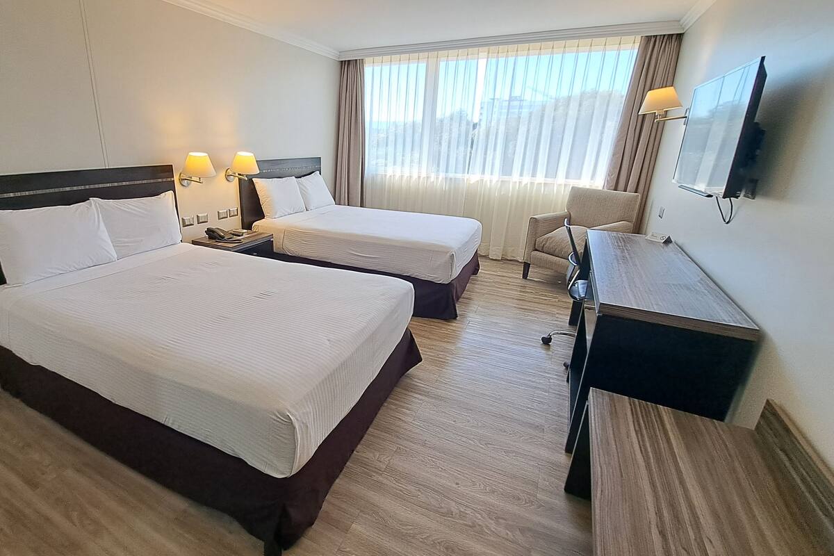 A hotel room with two beds, a desk, a chair, a television, and a window with curtains.