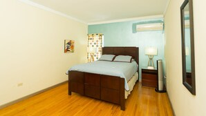 Une chambre avec un plancher en bois, un lit avec une tête de lit, une table de chevet, une lampe et un miroir.