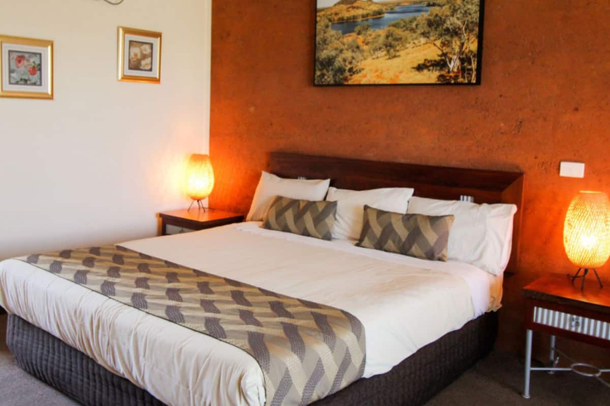 A neatly made bed with a patterned bedspread, two bedside lamps, and a painting of a landscape on the wall.