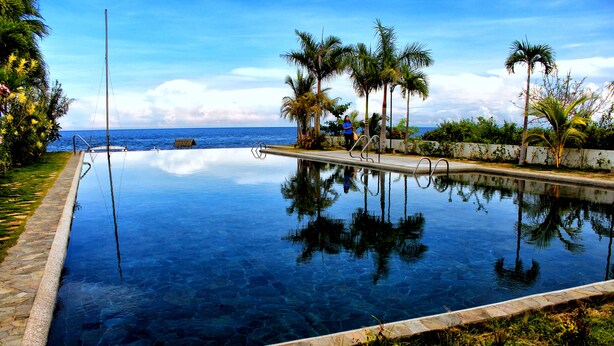 An infinity pool with clear water, surrounded by palm trees and a stone border, reflecting the sky and palm trees. - Bravo Resorts - Munting Paraiso (Dauin)