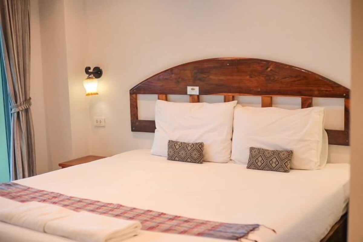 A neatly made bed with white linens and two patterned pillows, a wooden headboard, and a wall-mounted light fixture.