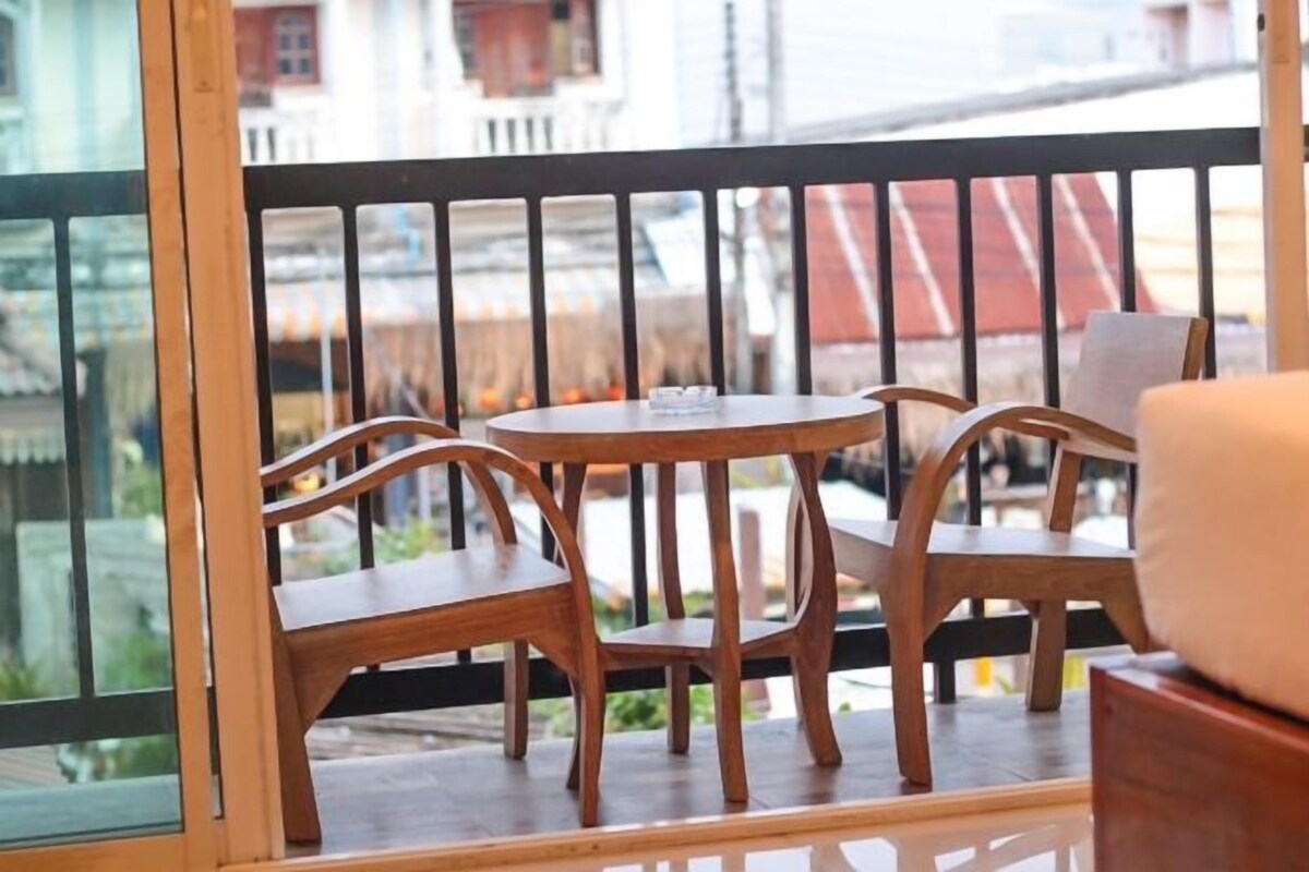 Un balcón con mobiliario de madera y barandilla de cristal, con vista a una calle con edificios.