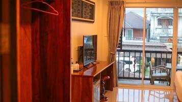 A room with a wooden desk, a television, and a balcony with a view of a traditional house and parked cars.