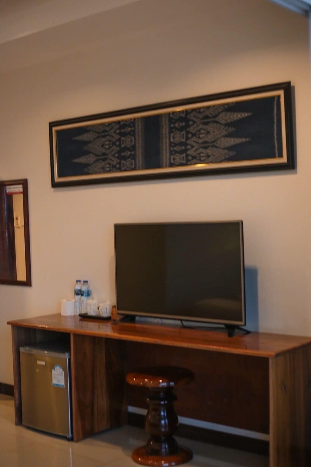 A hotel room with a wooden desk, a flat-screen TV, a small refrigerator, and a framed artwork above the TV.