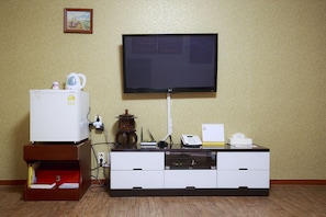 A room with a flat-screen TV mounted on the wall, a white refrigerator, a wooden cabinet with drawers, a small wooden table with a lamp, and a calendar on the cabinet.