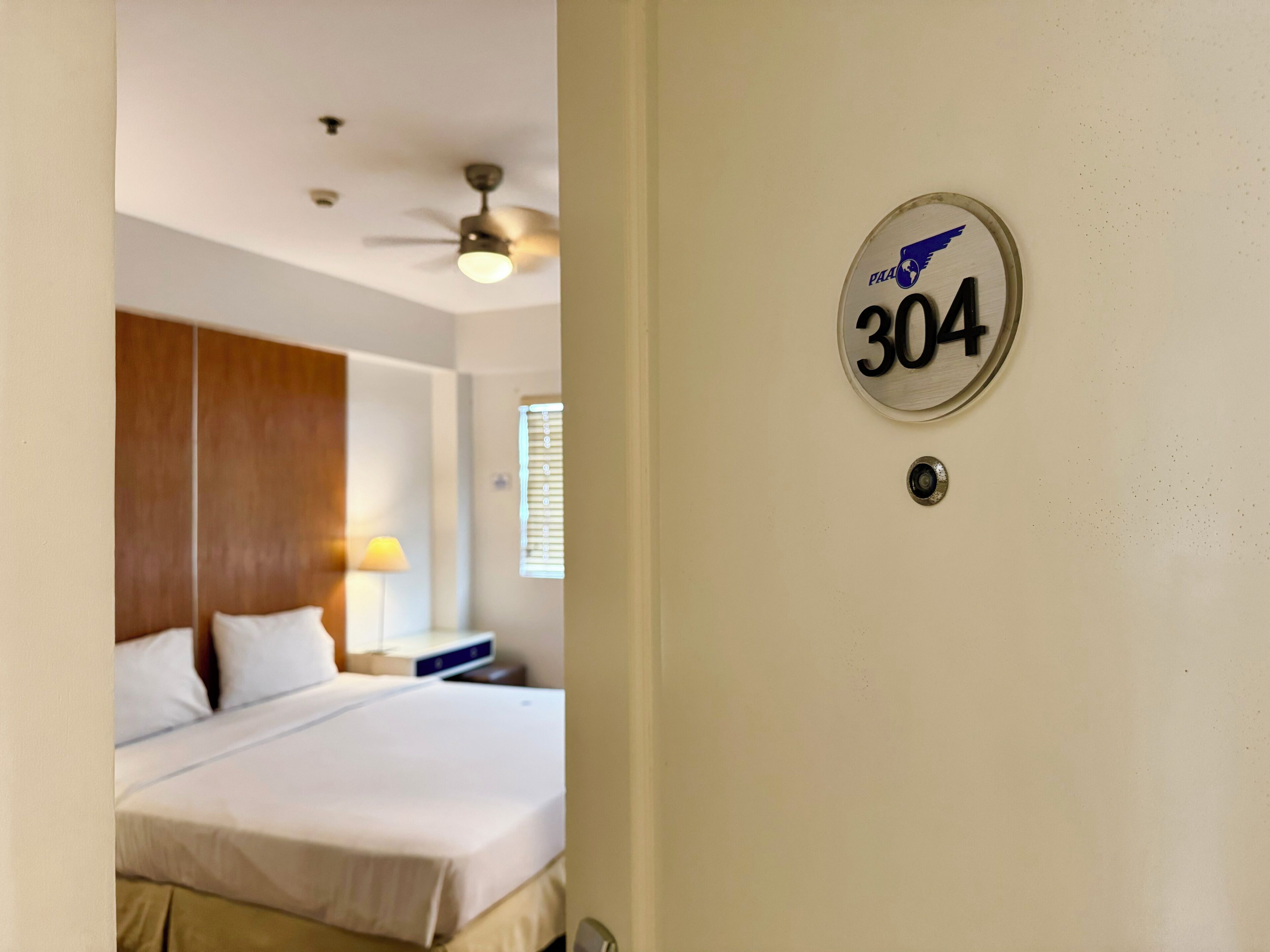 A hotel room with a bed, a ceiling fan, and a wall-mounted number plate.