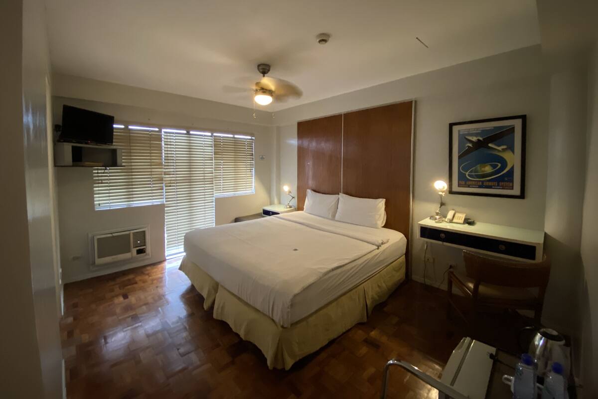 A hotel room with a large bed, a ceiling fan, a mounted TV, a desk with a chair, and a framed picture on the wall.